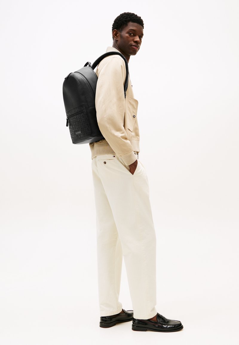 Zaino in pelle nera con tasca frontale texture, indossato con un maglione beige e pantaloni color crema, caratterizzato da una silhouette rilassata.