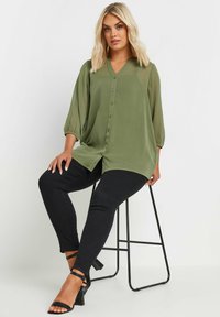 Blusa verde oliva a maniche lunghe con scollatura a V, chiusura con bottoni e maniche plissettate. Indossata con pantaloni neri aderenti e tacchi aperti.
