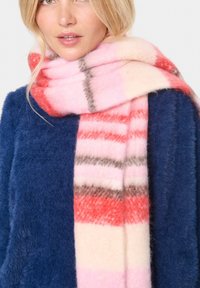 Bufanda de lana con rayas alternadas en rosa, rojo y gris, con una textura suave y esponjosa. Llevada sobre un suéter azul marino.