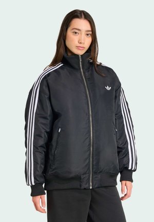 Young woman with long hair wears a black bomber jacket with white stripes and stands against a light background.