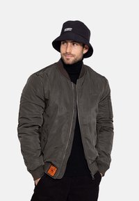 Olivegrüne Bomberjacke mit gerippten Bündchen, Reißverschluss und seitlichen Taschen; kombiniert mit einem schwarzen Bucket Hat mit weißem Logo.
