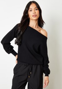Zwarte off-shoulder trui met lange, opgeblazen mouwen. Zachte textuur, geribbelde manchetten en een relaxte pasvorm. Gecombineerd met zwarte broeken met elastische taille.