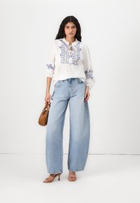 Blouse blanche avec broderie florale bleue, col à volants, associée à un jean large bleu clair, tenant un petit sac à main marron, portant des talons beiges.