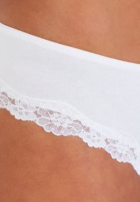 Witte katoenen onderbroek met een gebogen kantafwerking aan de bovenkant, voorzien van een bloemenpatroon langs de rand. Zachte textuur, naadloos ontwerp.