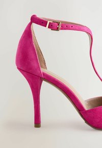 Zapato de tacón alto de ante rosa con punta afilada, tira en el tobillo con una hebilla en tono dorado y tacón de aguja delgado.