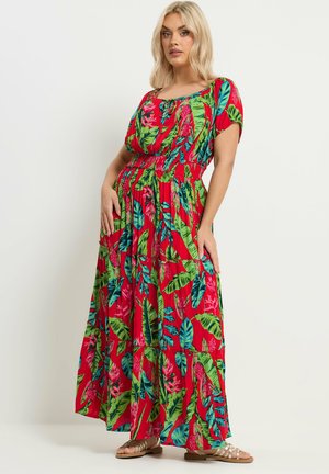 Lyshåret kvinde iført en rød maxi-kjole med grønne og blå tropiske bladprint, stående med hænderne på hofterne mod en ensfarvet baggrund.
