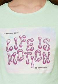 Hellgrünes Baumwoll-T-Shirt mit einem großen Grafikdruck des Satzes "LIFE IS MOTION" in Pink, mit einem glänzenden, fließenden Design.