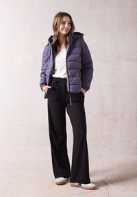 Giacca a vento viola con cappuccio, tasche con zip; T-shirt bianca; pantaloni neri a gamba larga; scarpe bianche con suole marroni e accenti neri.