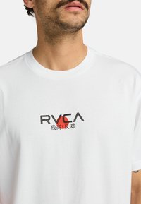 Camiseta de algodón blanca con un logotipo gráfico negro y rojo en el centro, con caracteres japoneses debajo. Corte regular.