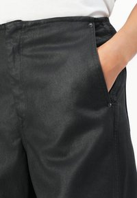 Gros plan sur un pantalon noir avec une main dans la poche avant droite, montrant la texture du tissu et les rivets en métal près des bords de la poche.