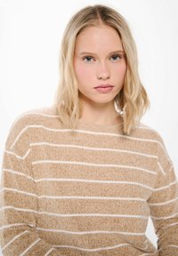 Beige randig stickad tröja med vita horisontella linjer, mjuk textur, lös passform och rund halsringning.
