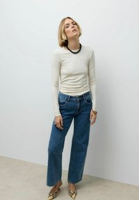 Vrouw met kort blond haar, gekleed in een crème lange mouwen top, blauwe wijde jeans, een zwarte kralenketting en slangenprint hakken, staande tegen een witte muur.