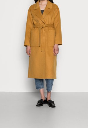 Person iført en sennepsgul kåbe med bælte og store lommer, kortede blå jeans og sorte skinnende slip-on sko, stående på gråt gulv.