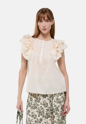 Femme portant un chemisier crème à manches volantées et boutons, associé à une jupe beige avec un motif floral foncé, tenant des bretelles assorties.