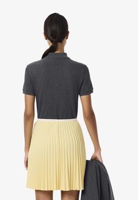 Grå kortärmad polo skjorta i kombination med en plisserad gul kjol med vit midja. Modellen håller en grå jacka.