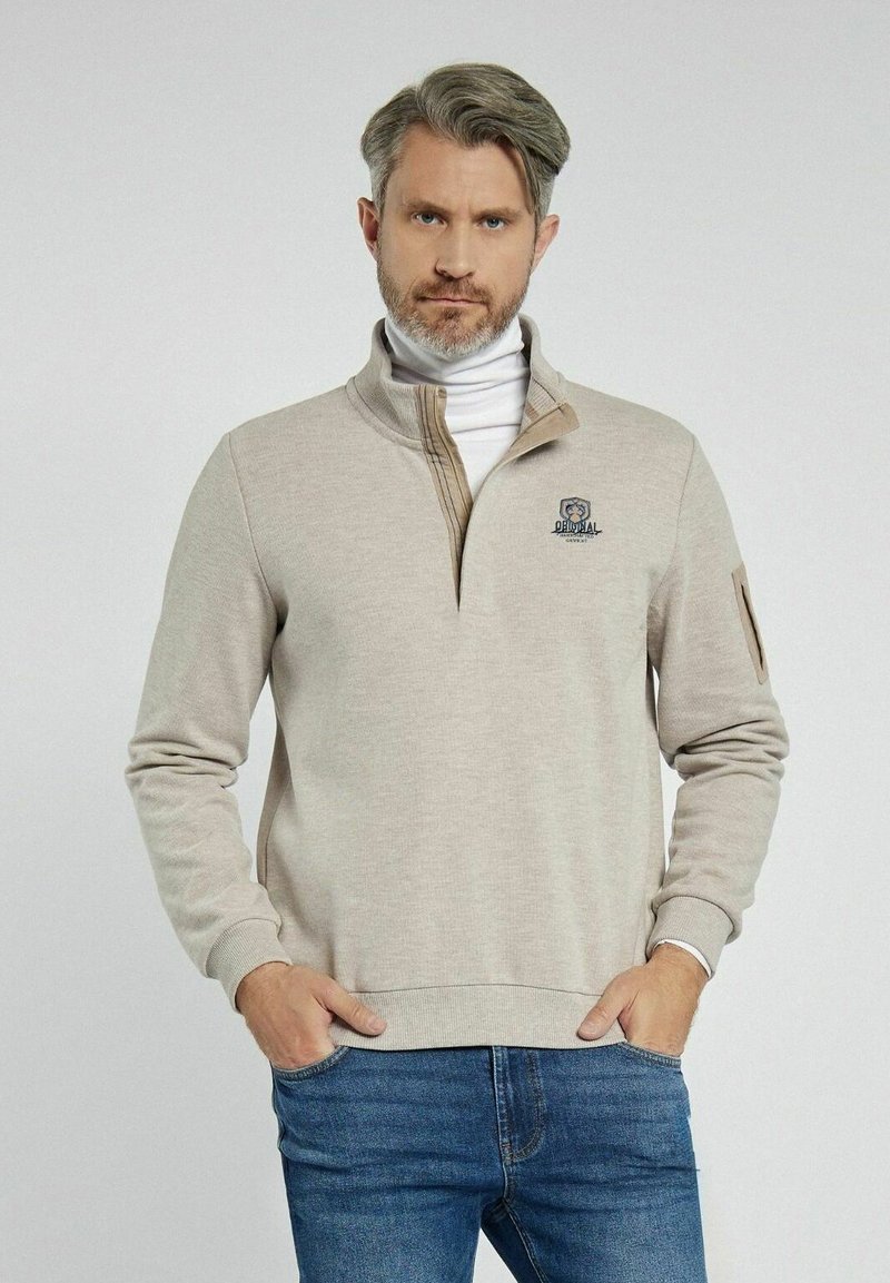 Beżowy sweter z kołnierzem na pół zamek, z haftowanym logo na piersi oraz kontrastowymi akcentami. Wykonany z miękkiego materiału, o luźnym kroju.