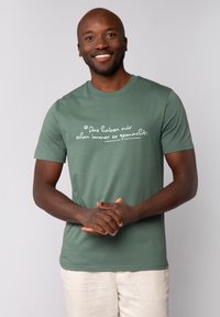 T-shirt en coton vert à manches courtes, avec du texte blanc dans une police décontractée sur la poitrine. Le modèle sourit, les mains jointes.