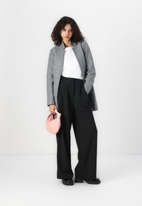 Grijze wollen blazer zonder kraag, witte blouse, zwarte wijde broek, en een kleine roze handtas. Zwarte stevige schoenen.