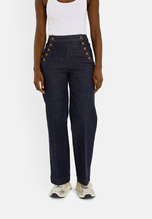 Pantalones azul oscuro de talle alto y pernera ancha con dos filas de botones en cada lado, llevados con una camiseta sin mangas blanca y zapatillas de color claro.