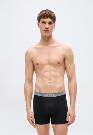 Νέος άνδρας όρθιος, γυμνός στο στήθος, φορώντας μαύρα εσώρουχα boxer Levi’s με γκρι μέση, μπροστά σε απλό φωτεινό φόντο.