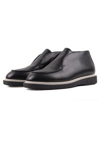 Chaussures noires en cuir à enfiler avec bouts arrondis, liseré blanc et semelles en caoutchouc noir texturé, présentées côte à côte sur fond blanc.