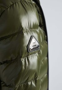 Olijfgroene pufferjack met glanzende afwerking, voorzien van een driehoekige zwarte logopatch en gevoerde secties voor isolatie. Ritsdetail zichtbaar.