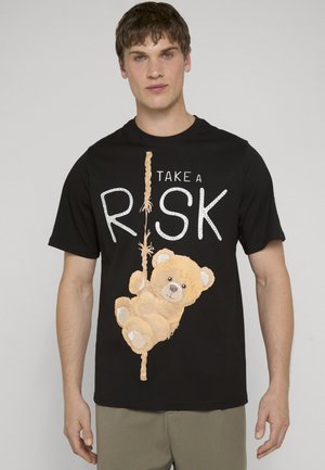 Młody mężczyzna w czarnej koszulce z misiem wspinającym się po linie i napisem „TAKE A RISK” z przodu.