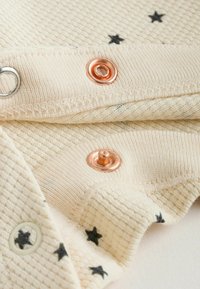 Gros plan sur un tissu beige avec des imprimés d'étoiles noires et des boutons-pression couleur cuivre sur un tricot texturé.