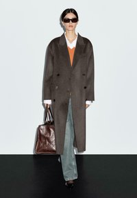 Manteau croisé en mélange de laine marron foncé, pull orange, chemise blanche, jeans bleus, chaussures noires à bouts pointus et sac fourre-tout en cuir marron.
