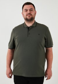 Olívovo zelená polokošeľa s krátkymi rukávmi, rebrovaným golierom, zapínaním na tri gombíky a malým logom na hrudi. Hladká textúra látky.