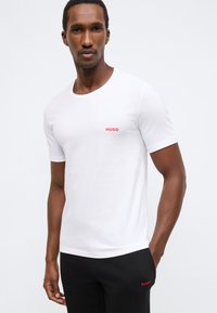 Balta trumpomis rankovėmis medvilninė marškinėliai su apvalia apykakle; kairėje krūtinės pusėje yra mažas raudonas „HUGO" logotipas.