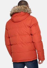 Homme portant une veste matelassée orange avec une capuche en fausse fourrure, vu de dos, associée à un jean bleu sur un fond clair uni.