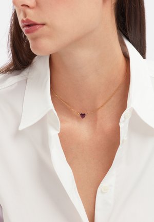 Gouden ketting met een hartvormige paarse edelsteen en kleine gouden accenten, gedragen over een wit collshirt.