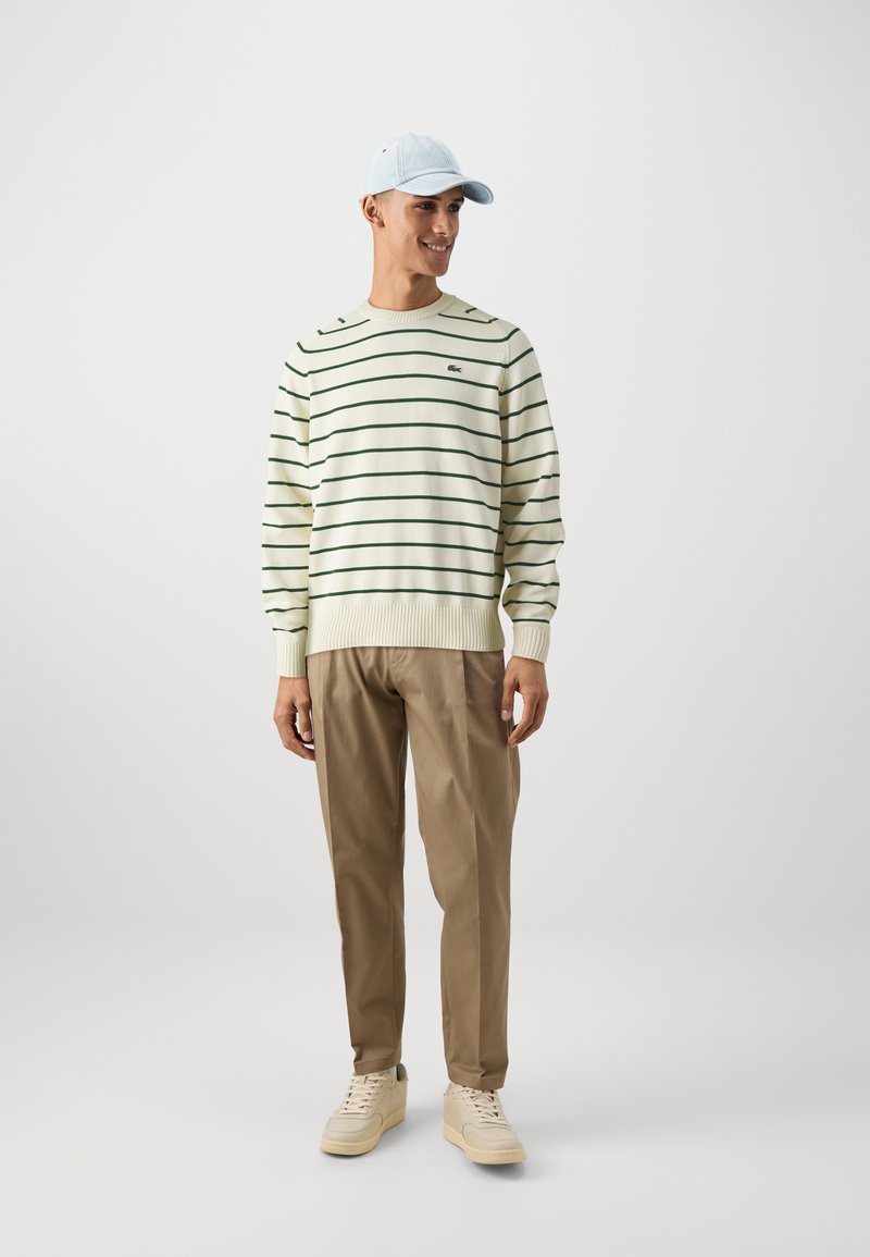 Suéter crema a rayas con líneas verdes, pantalones beige y gorra azul claro, combinados con zapatillas beige. Presenta un ajuste relajado y detalles acanalados.