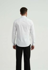 Chemise blanche à manches longues avec un col boutonné, présentant des poignets contrastés et un ourlet arrondi, portée avec un pantalon noir, vue de dos.