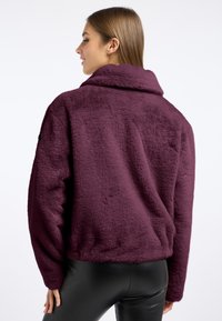 Fioletowa kurtka z sztucznego futra o miękkiej teksturze, z szerokim kołnierzem i opadłymi ramionami. Posiada prążkowane mankiety i luźny krój.