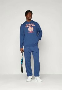 Ναυτικό μπλε φούτερ με γραφική εκτύπωση που αναγράφει "Stockholm Monte Carlo Tennis Club", συνδυασμένο με ταιριαστά παντελόνια φόρμας και λευκά αθλητικά παπούτσια, κρατώντας ένα τένις ρακέτα.