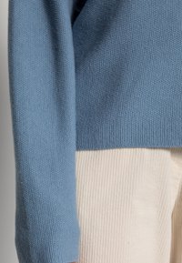 Niebieski sweter z dzianiny na rękawie i dolnej krawędzi, noszony na boku tułowia i ramieniu osoby, zestawiony z beżowymi spodniami z prążkowanej korduroy.