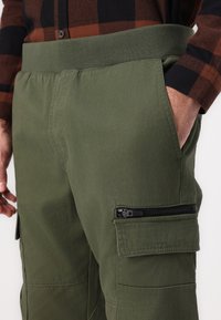 Olivgrön cargobyxor med elastisk midja, sidofickor och en zip-ficka på låret. Tillverkade av slitstarkt tyg med en mjuk yta.