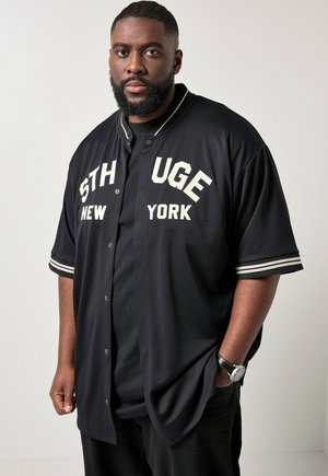 Sort mand med skæg iført en sort varsity-stil jakke med hvid tekst "STHUGE NEW YORK" og et sort ur, stående med den ene hånd i lommen.