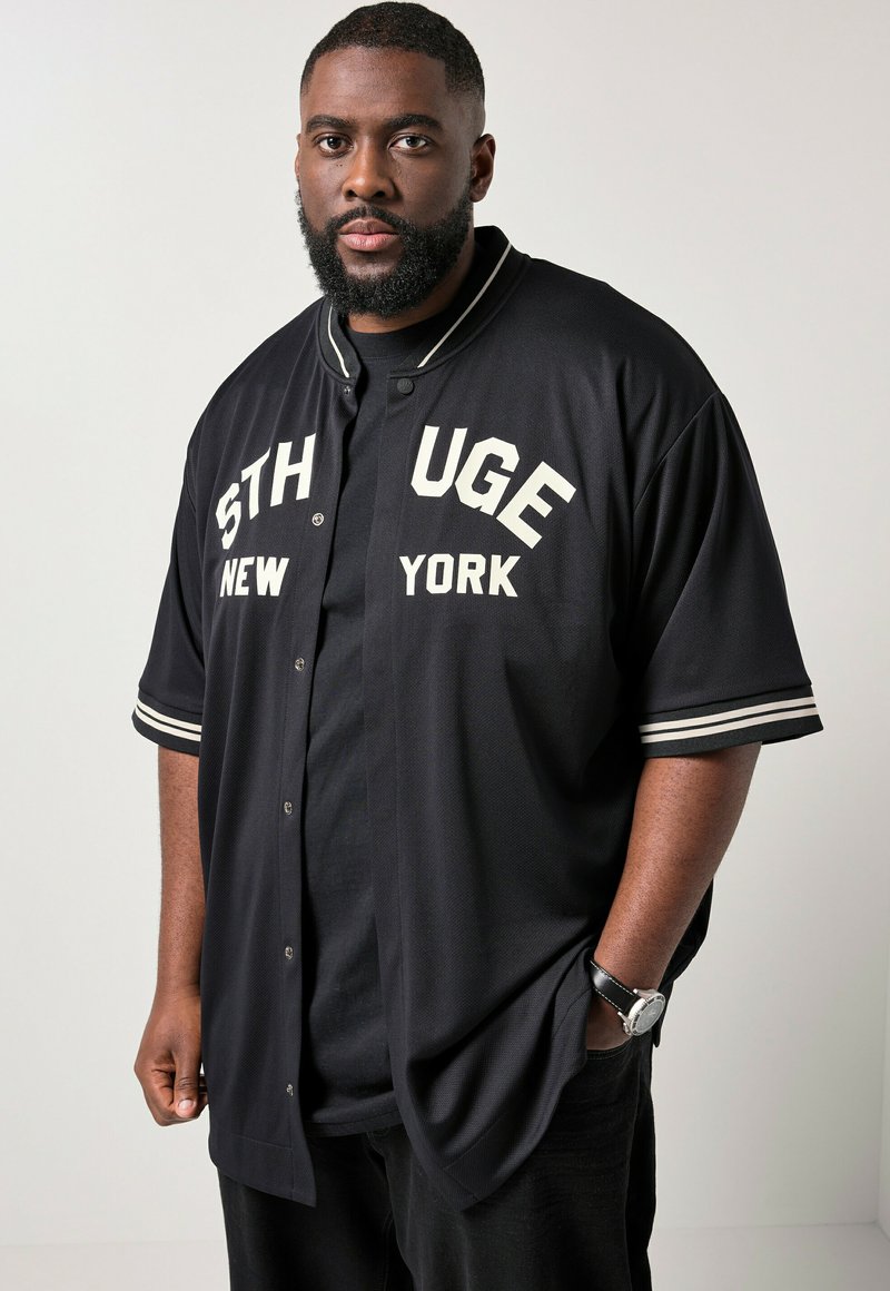 Schwarzer Mann mit Bart, der eine schwarze Varsity-Jacke mit weißem Schriftzug "STHUGE NEW YORK" und eine schwarze Uhr trägt, steht mit einer Hand in der Tasche.
