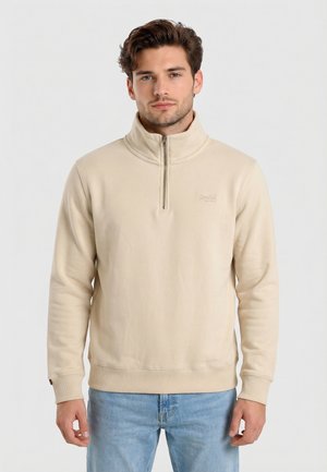 Giovane uomo con capelli castani che indossa una felpa beige con cerniera a quarto e jeans azzurri, in piedi davanti a uno sfondo chiaro e uniforme.