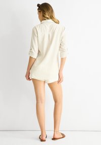 Witte, gestructureerde shirt met opgerolde mouwen, detail met rugplooi en een relaxte pasvorm. Gekocht met witte shorts en bruine platte sandalen.