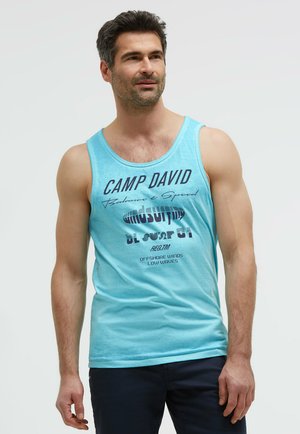 Man wearing a light blue sleeveless shirt with "Camp David" text, standing with a slight smile against a plain background.
