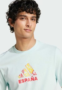 Camiseta de algodón azul claro con el texto "España" en rojo y un logo de Adidas multicolor con patrones geométricos. Cuello redondo y mangas cortas.