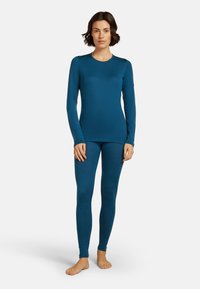 Woman standing barefoot wearing a fitted, long-sleeve blue thermal top and matching leggings against a plain white background.