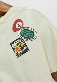 Heledbeez t-särk, millel on kolm värvilist plaastrit: punane ring logoga, roheline globuse embleem ja must "BANG!" graafika tähtedega.