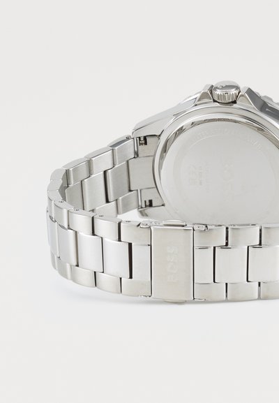 Montre en acier inoxydable argenté avec un bracelet à maillons poli, présentant un boîtier rond, une couronne texturée et un logo de la marque gravé sur le fermoir.