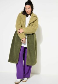 Cappotto lungo oversize di un verde oliva, con texture e un esterno similpelliccia, ampio colletto e bottoni neri, abbinato a pantaloni larghi viola.