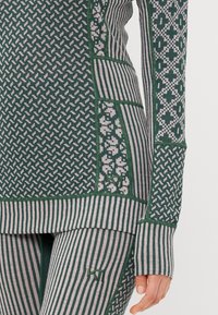 Zbliżenie na tors i ramię osoby ubranego w zielony i beżowy wzorzysty sweter oraz legginsy z geometrycznymi i paskowymi wzorami.
